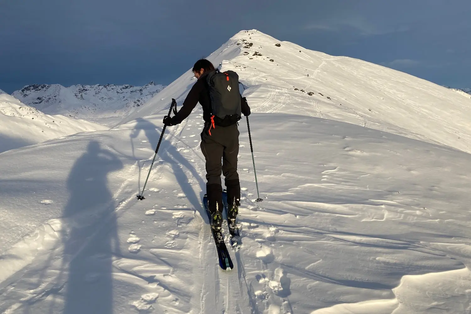 person wearing crique 25 backpack and ski gear is ascending a snowy mountain using ski poles