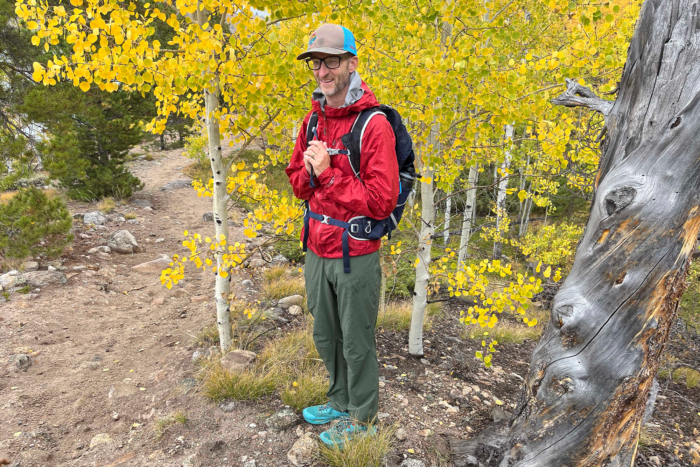 A man in Outdoor Research Ferrosi pants and a red rain jacket smiles while standing among yellow aspen trees