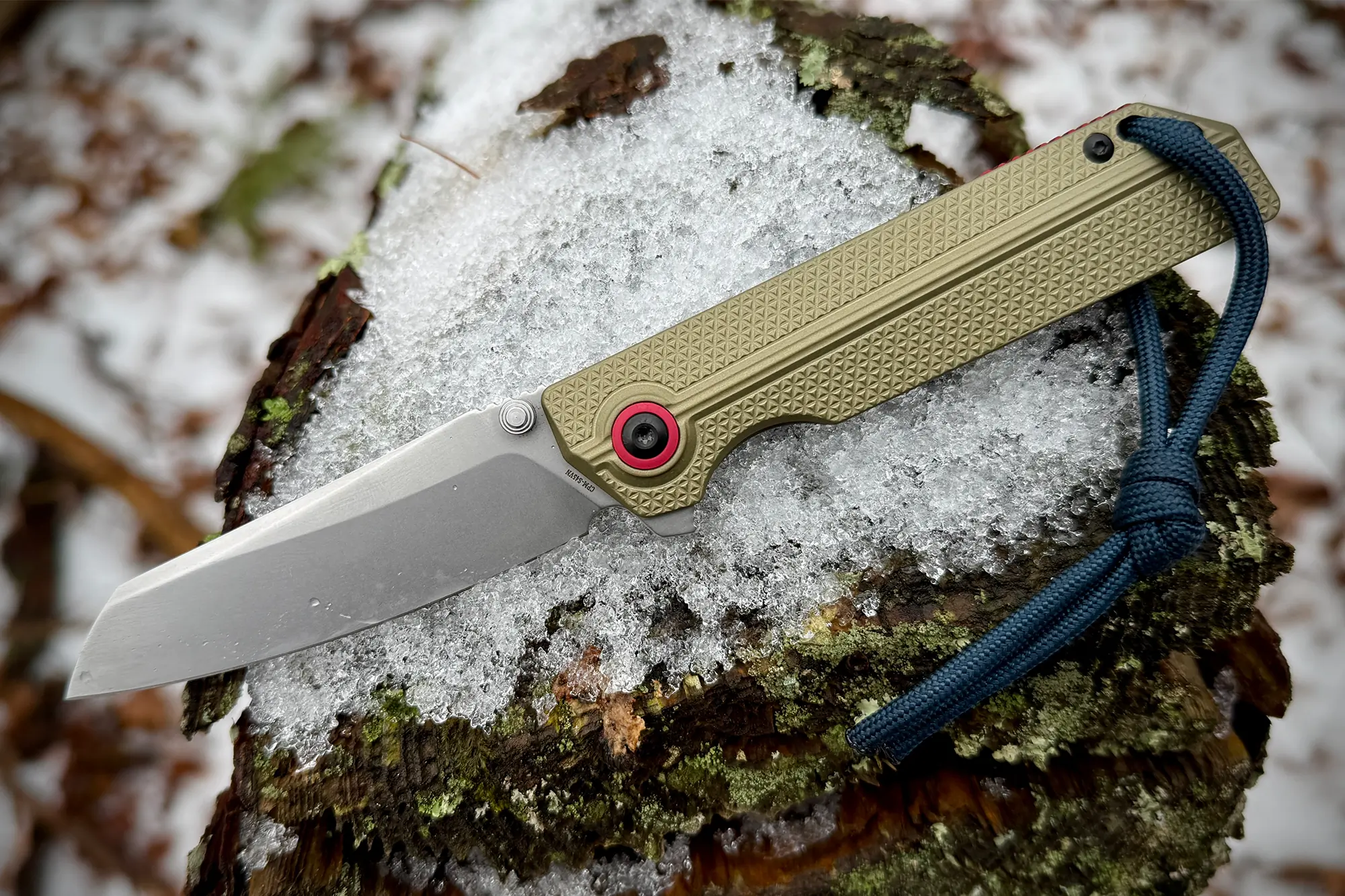 A side view of the OKNIFE XEN-OAL knife with an olive green textured handle and a silver blade, lying on a snow-covered tree stump