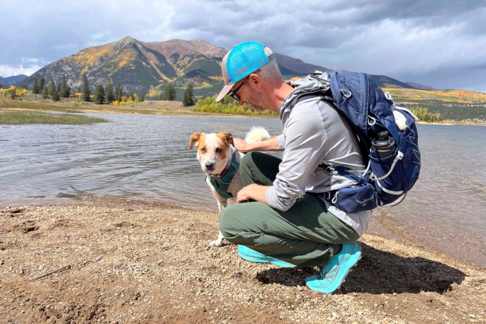 A man wearing Outdoor Research Ferrosi pants crouches beside a calm lake, petting his dog, with mountains in the background