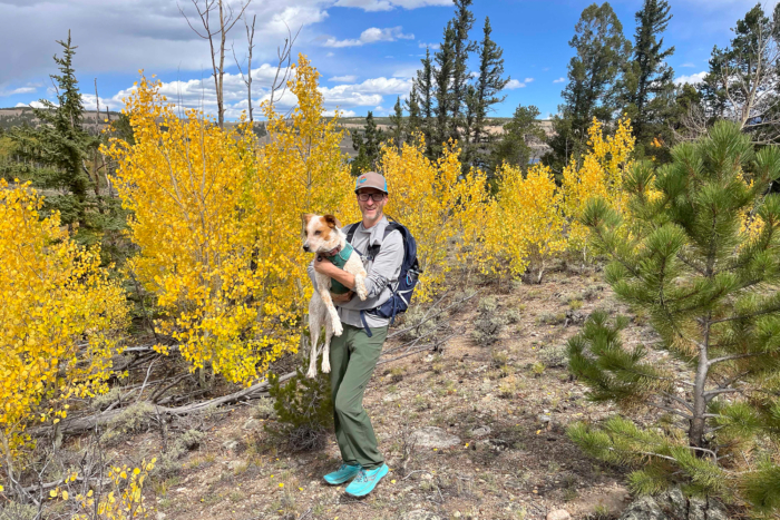 A hiker wearing green Outdoor Research Ferrosi pants with a backpack and turquoise shoes stands in a grove of bright yellow aspen trees