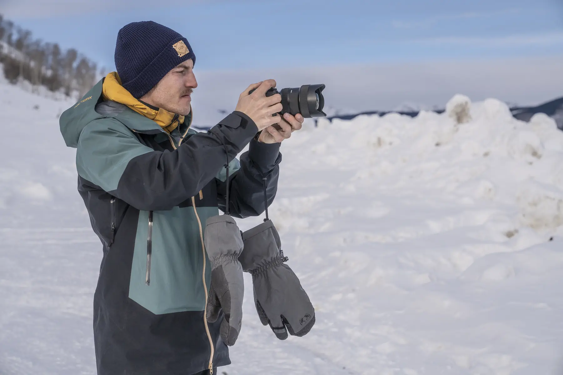 A man in a winter jacket takes photos, with Kuiu Northstar Glomitt gloves hanging from his wrists