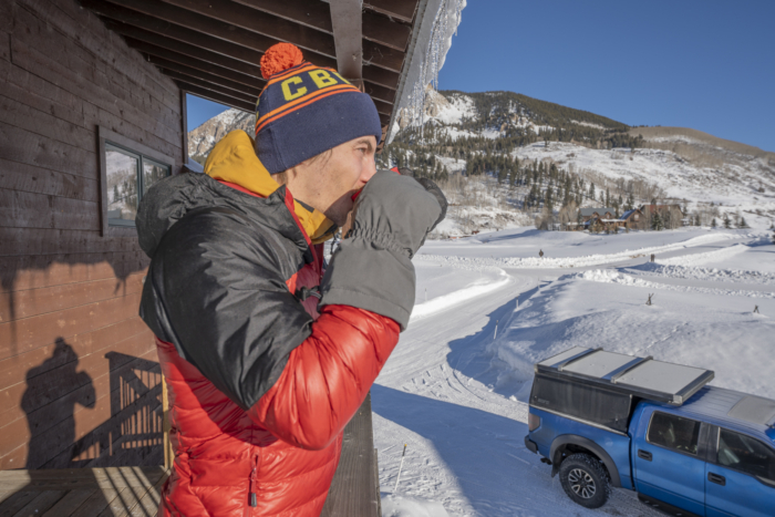 A man takes a sip from a red cup, wearing thick gloves and standing on a wooden balcony