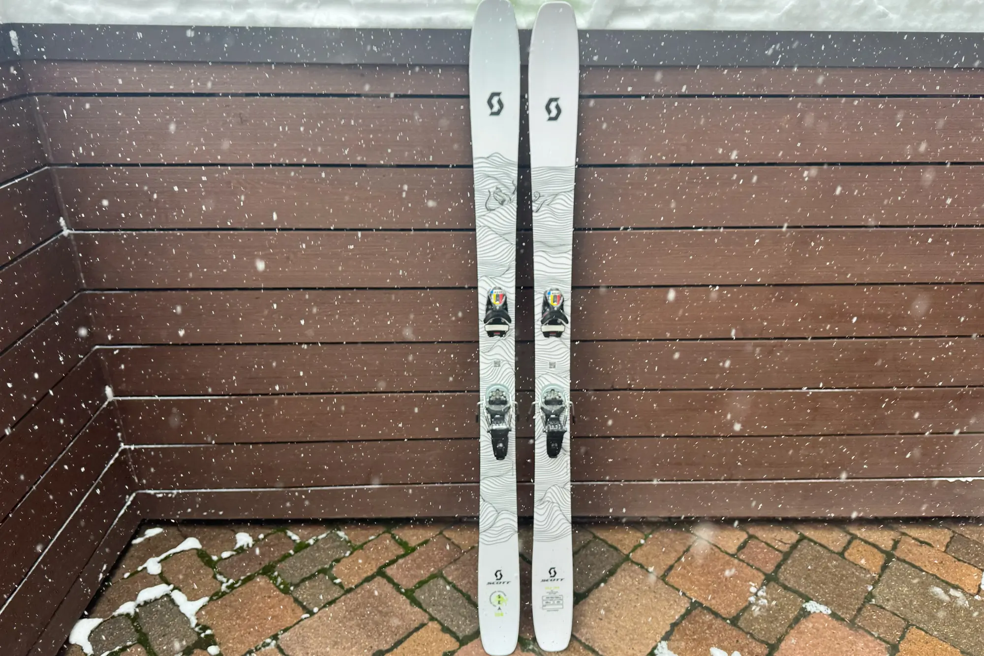 The Scott SEA 108 skis leaning against a wooden wall with snowfall