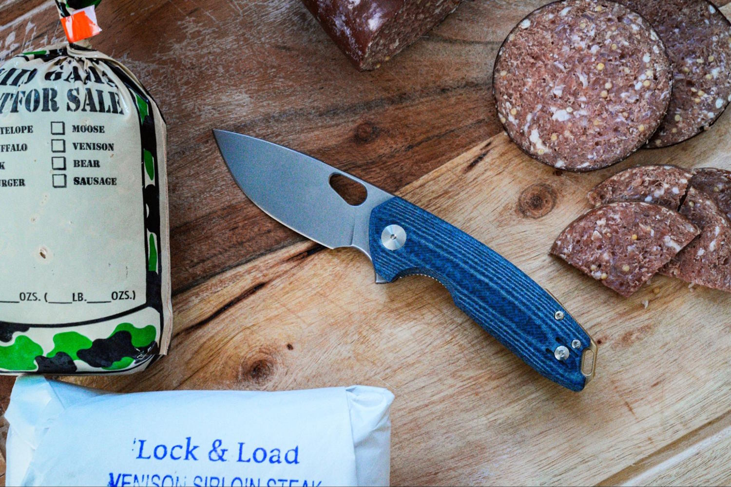 A GiantMouse ACE Tribeca knife lies on a wooden cutting board next to sliced sausage