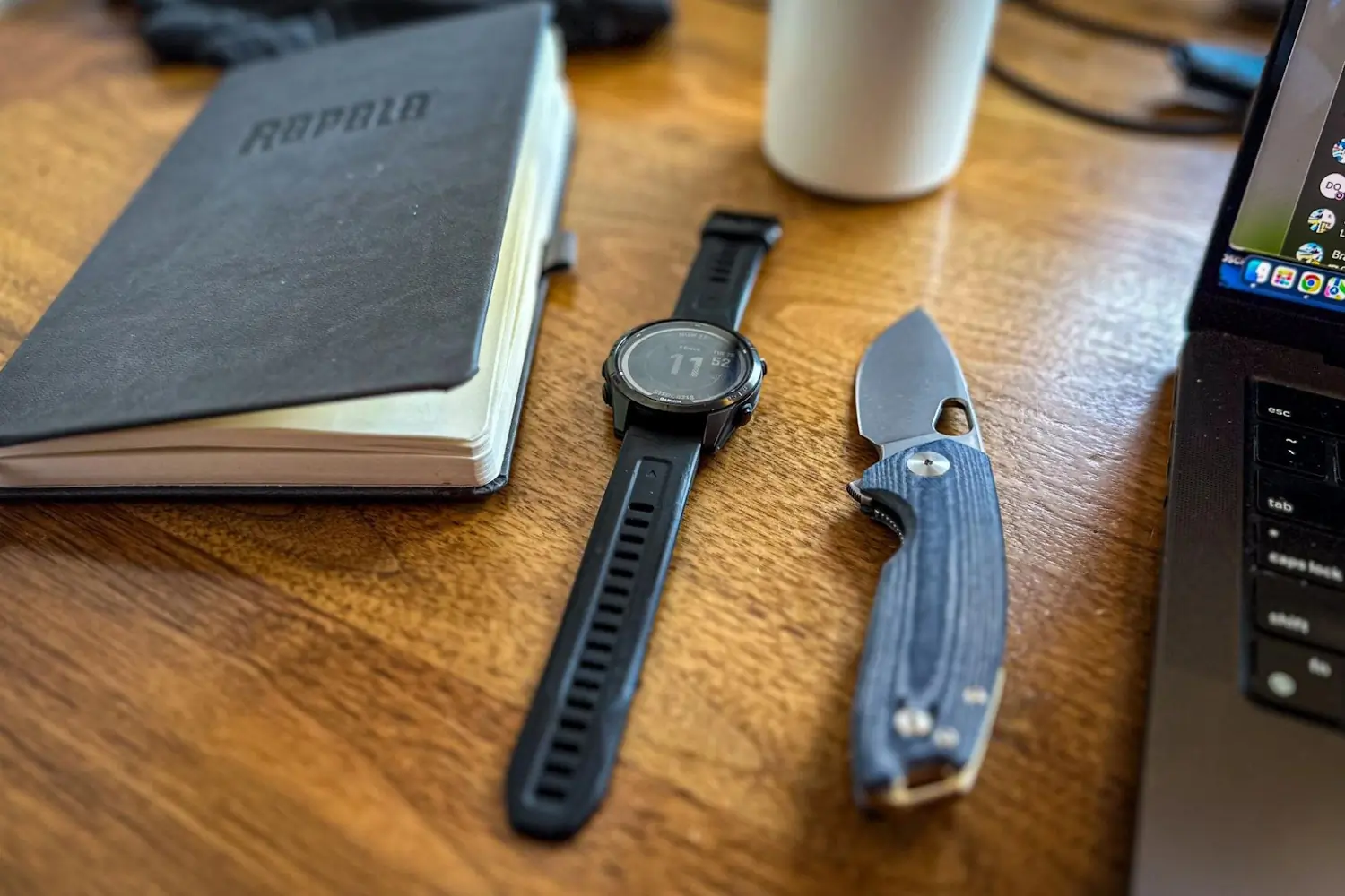 A blue-handled ACE Tribeca knife, a watch, and a notebook on a wooden desk