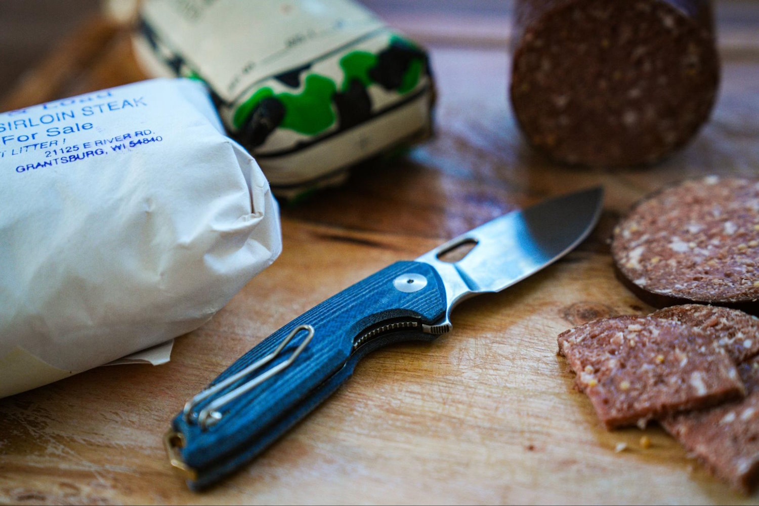 A GiantMouse ACE Tribeca knife lies on a wooden cutting board next to sliced sausage