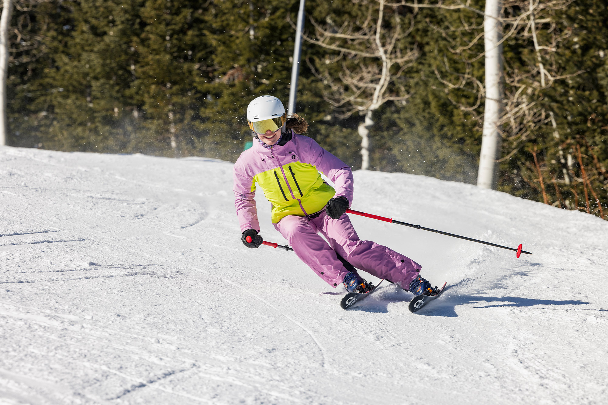 A skier in a bright pink and yellow jacket leaning into a turn on packed snow