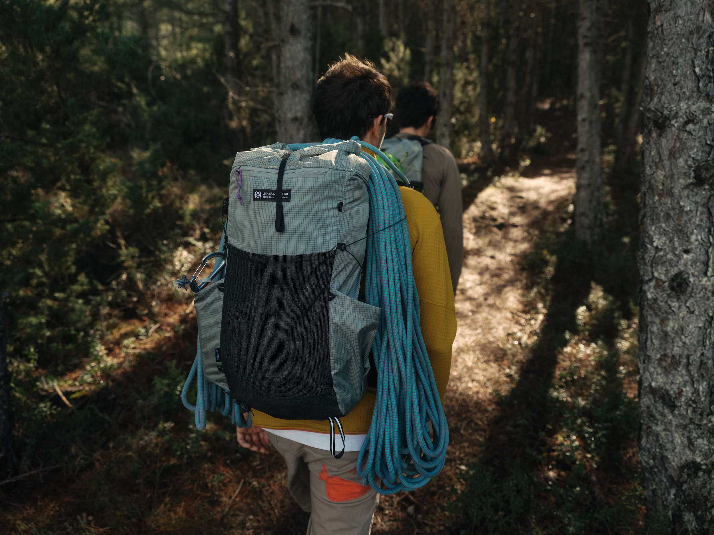 Two people walking through woods, one has a backpack with climbing rope over top.