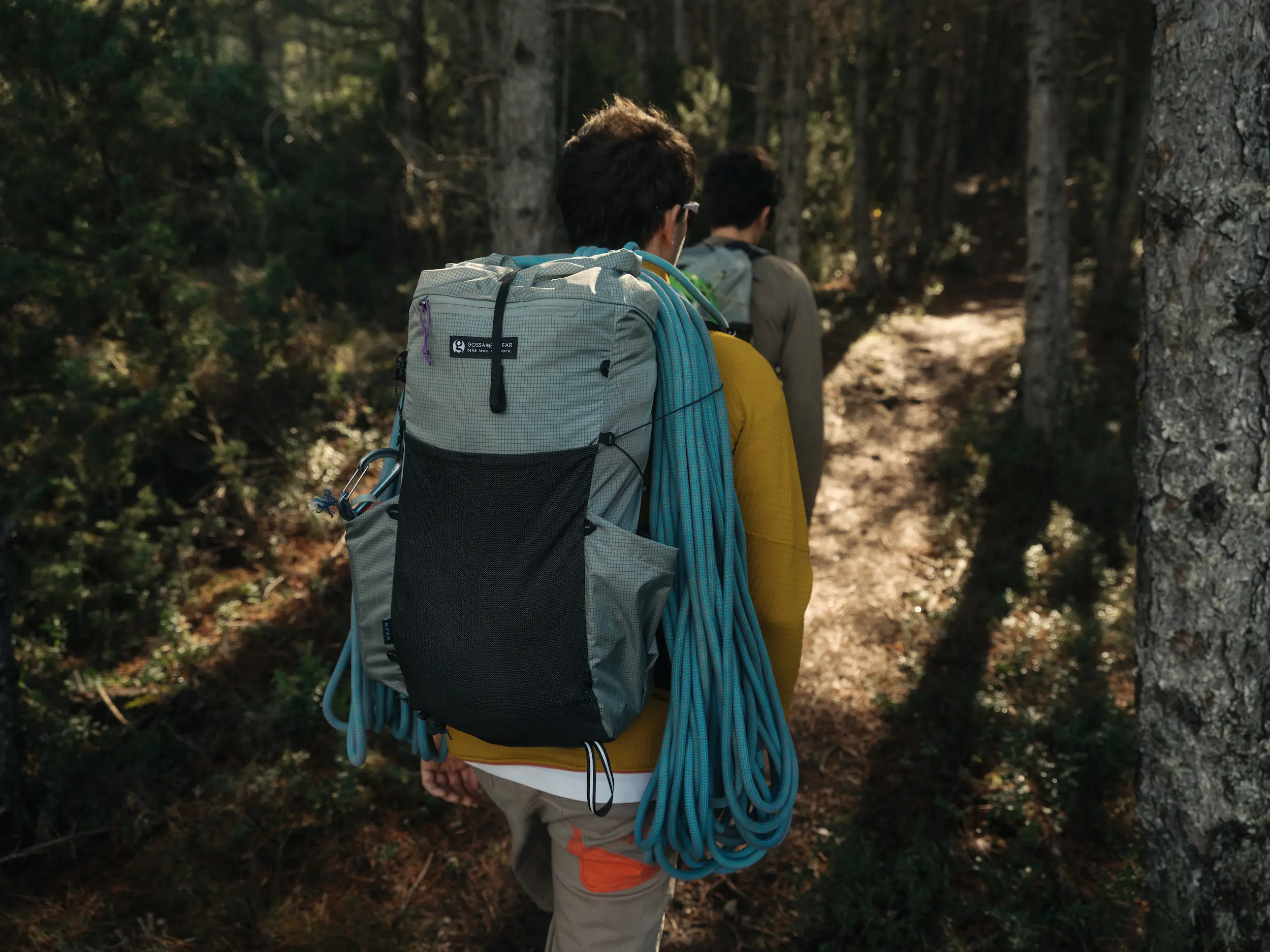 Two people walking through woods, one has a backpack with climbing rope over top.