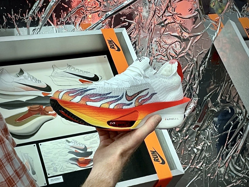 A hand holds a white Nike Alphafly shoe with flame details and a red-to-yellow gradient midsole in a design studio