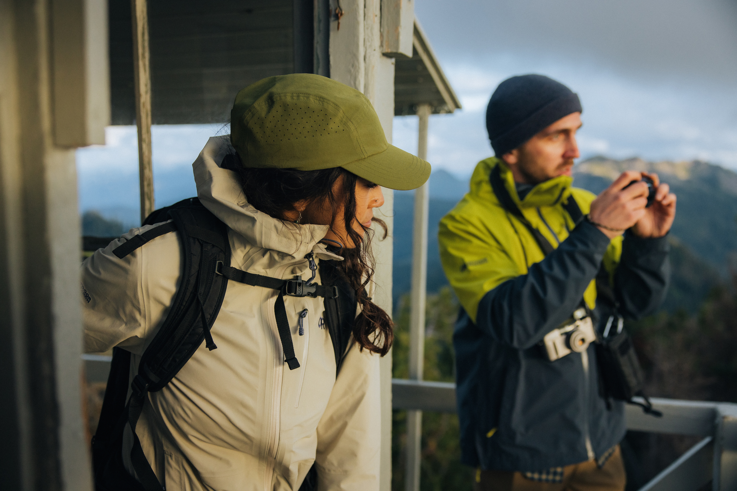 Man and woman in rain jackets on tower overlooking mountains.