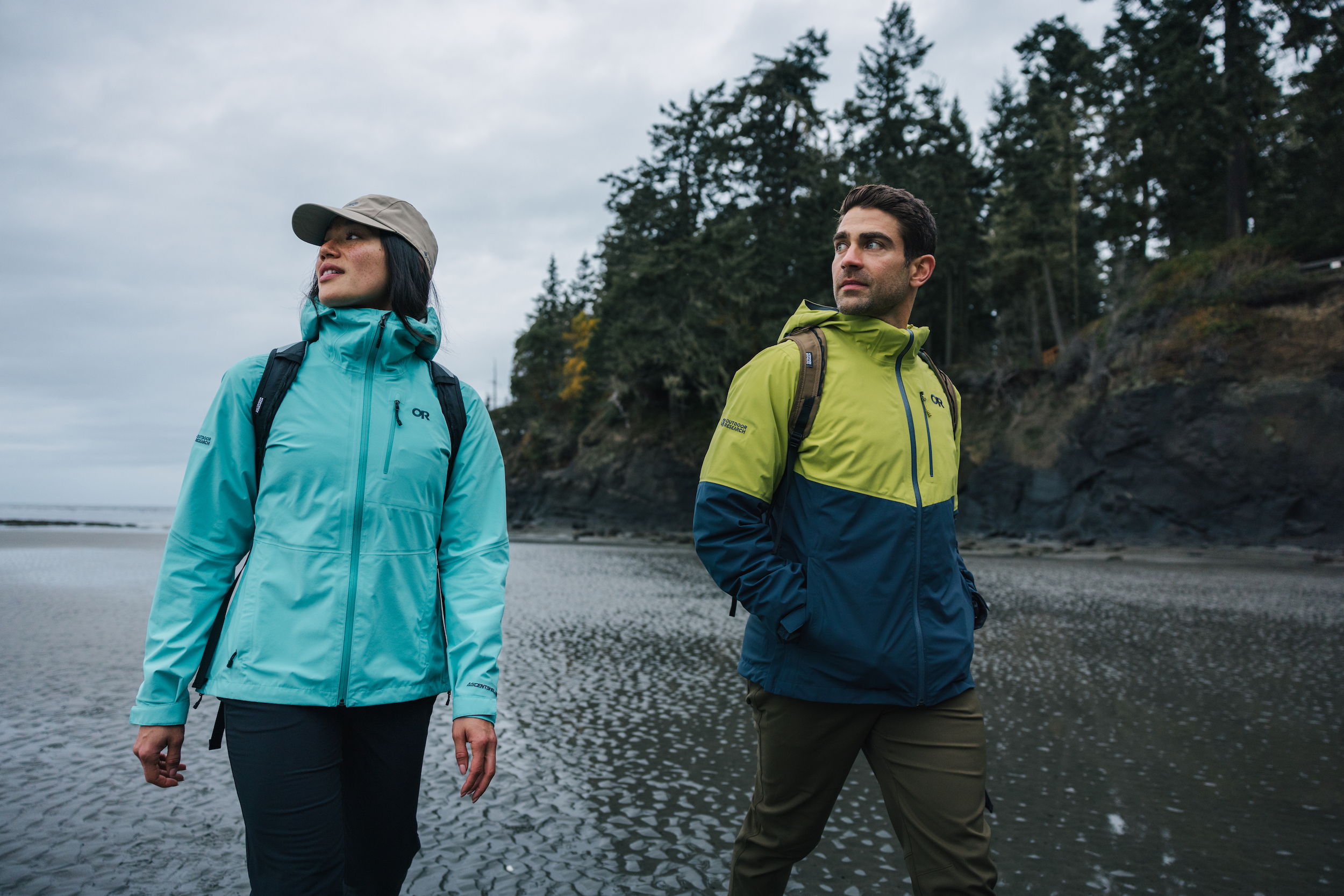 Woman and man in rain jackets and backpacks on beach with trees.