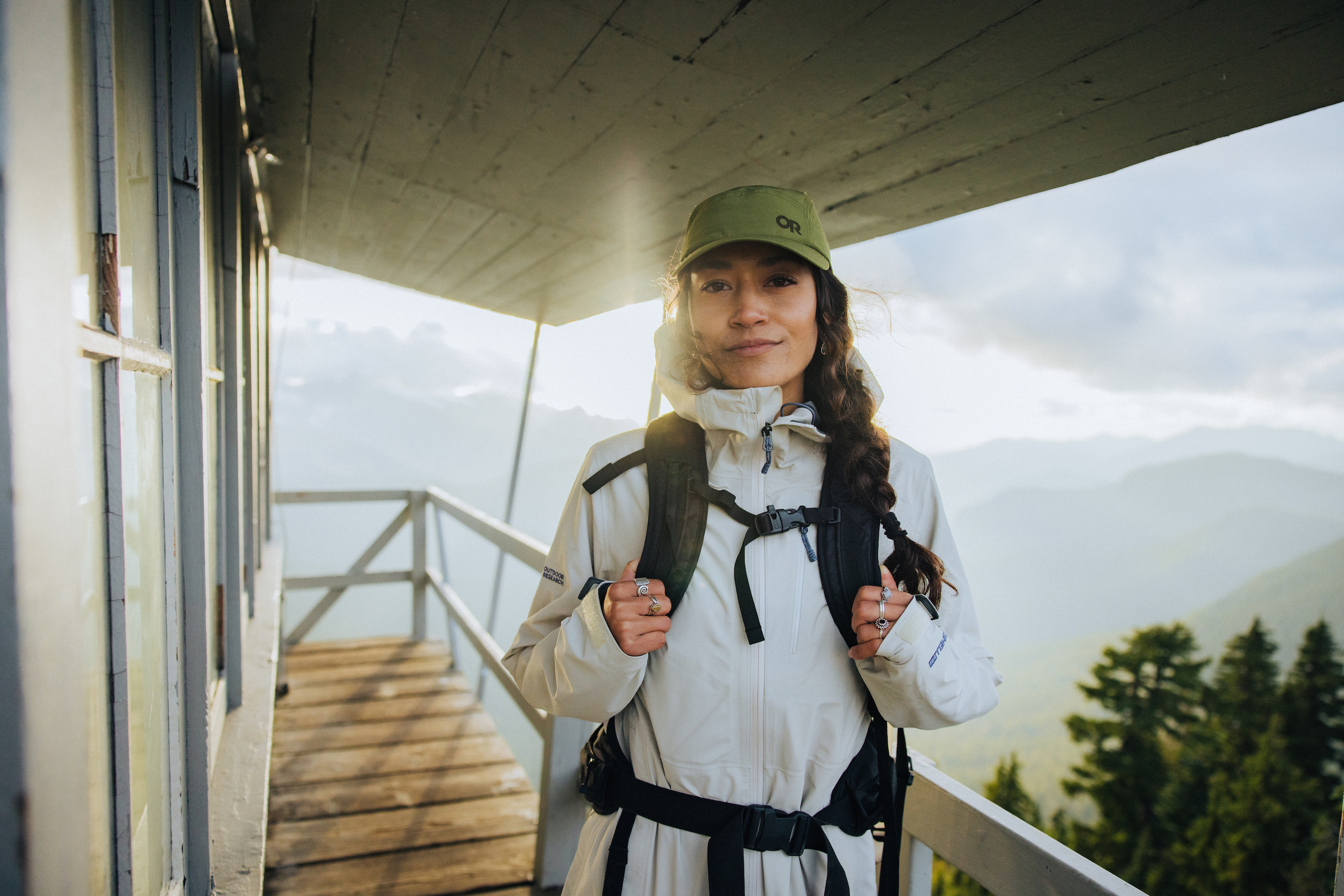 Woman wearing rain jacket, hat, and backpack on a fire tower with mountains and sun behind her.