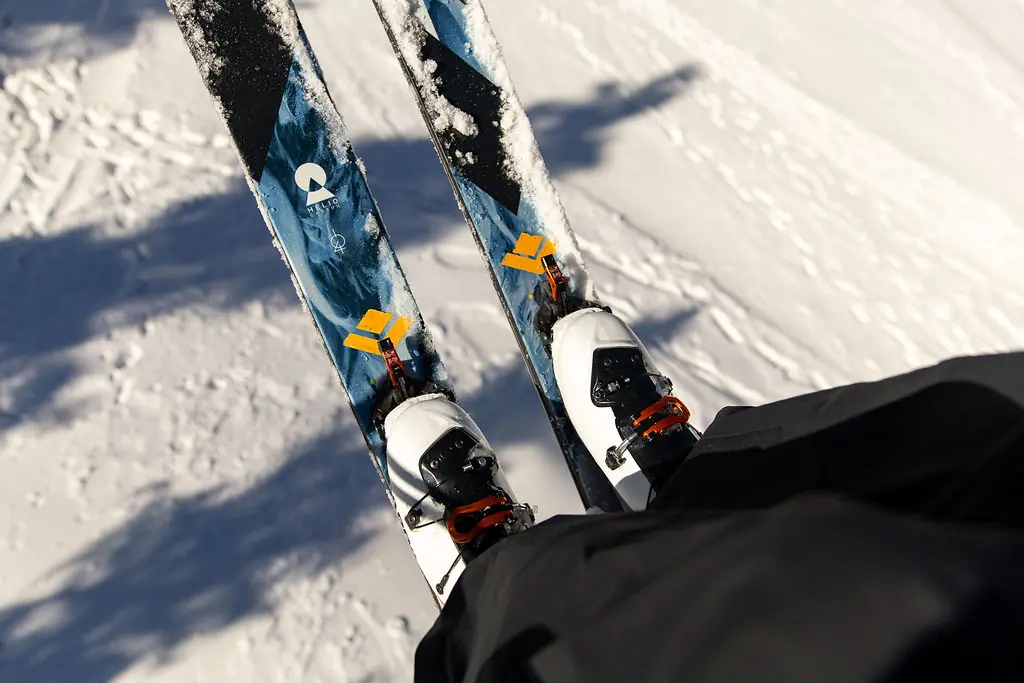 A skier looks down at their feet, showing the tops of blue and black skis with snow-covered bindings