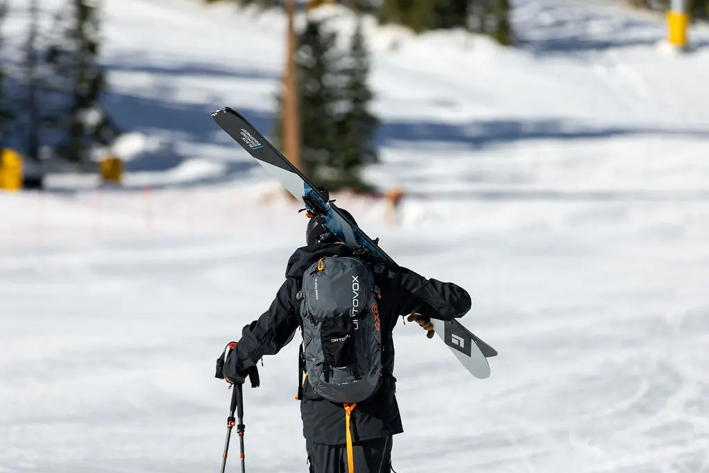 A skier in black gear carries skis equipped with ATK Raider 13 Evo bindings on their backpack while walking uphill on a snow-covered slope