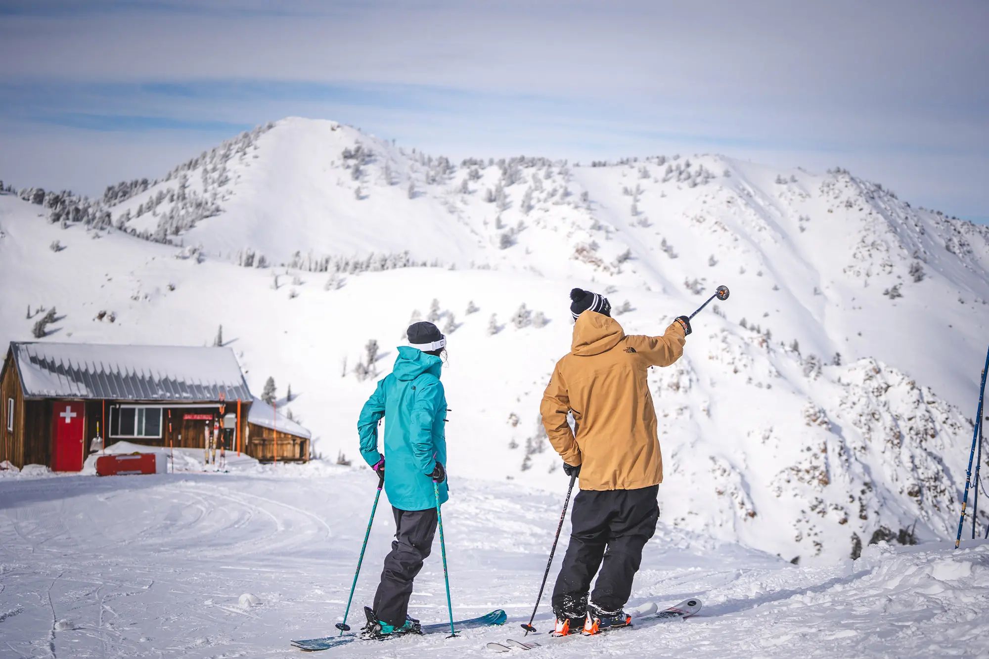 Two skiers stand near a ski patrol hut, looking at the slopes of Powder Mountain