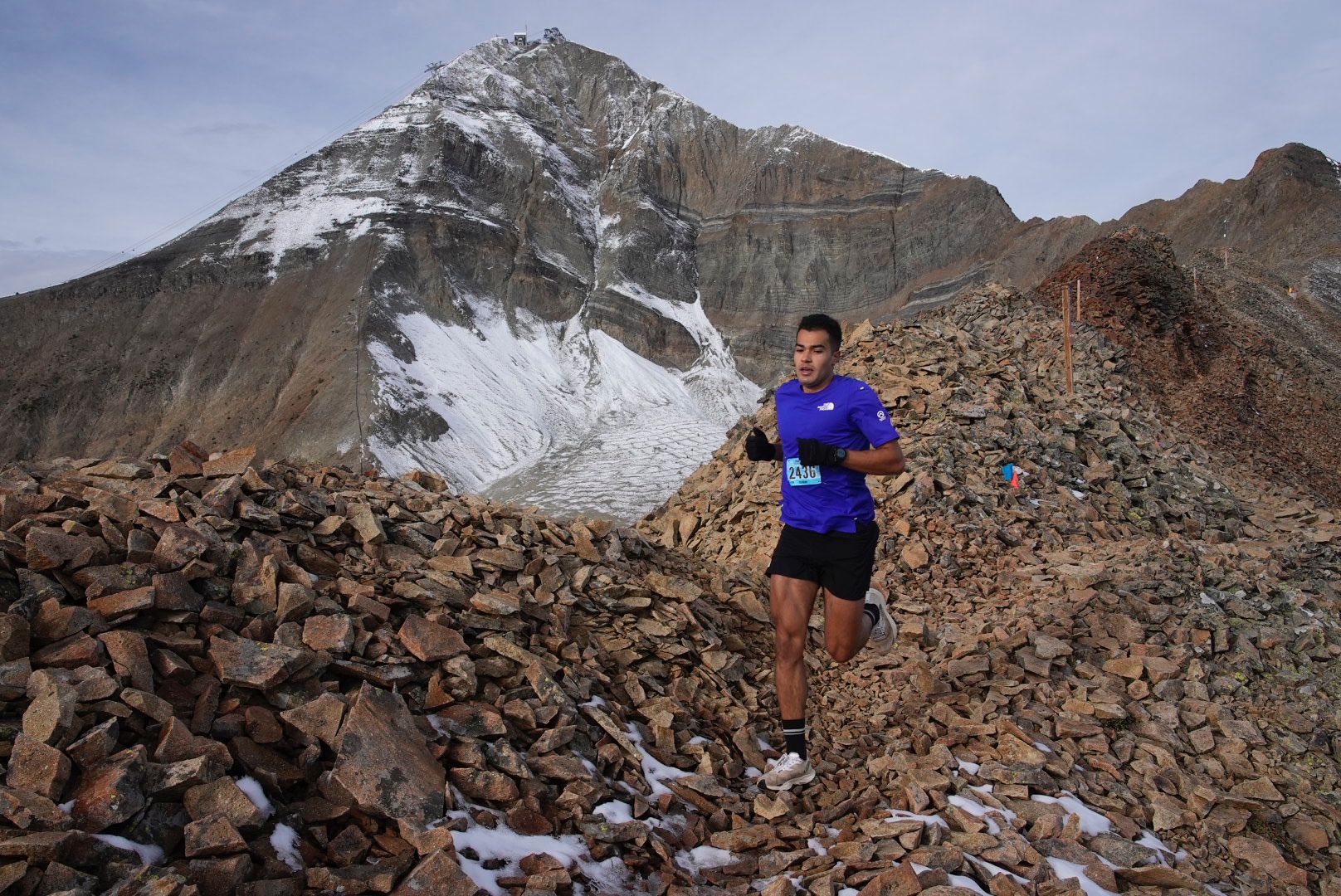 A trail runner in a blue shirt navigates rocky terrain high in the mountains, with a snow-covered peak in the background