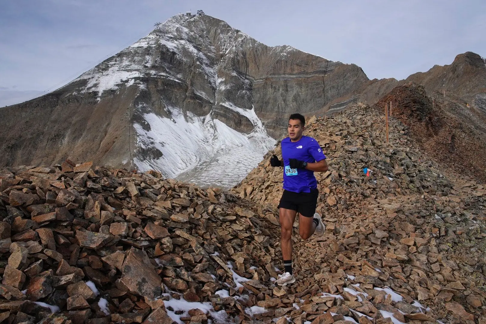 A trail runner in a blue shirt navigates rocky terrain high in the mountains, with a snow-covered peak in the background