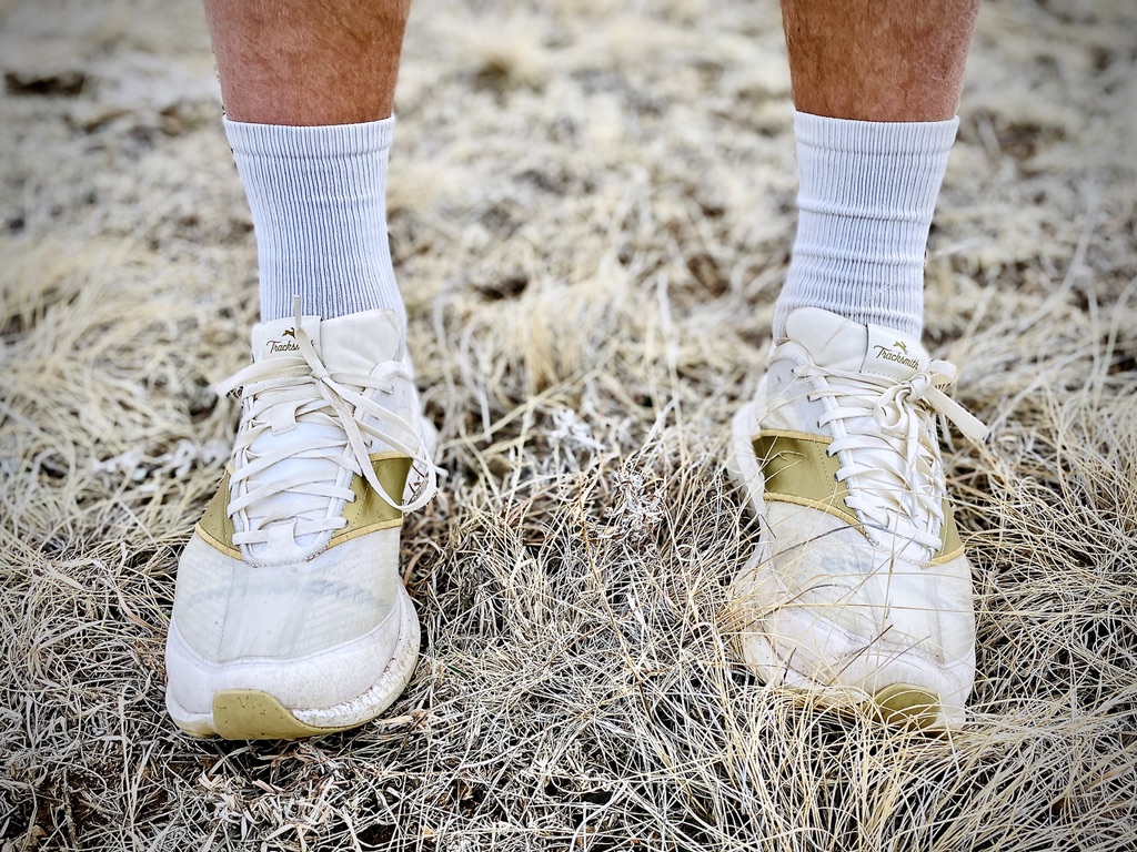 A pair of Tracksmith Eliot Racer shoes worn with white socks, standing on dry grass