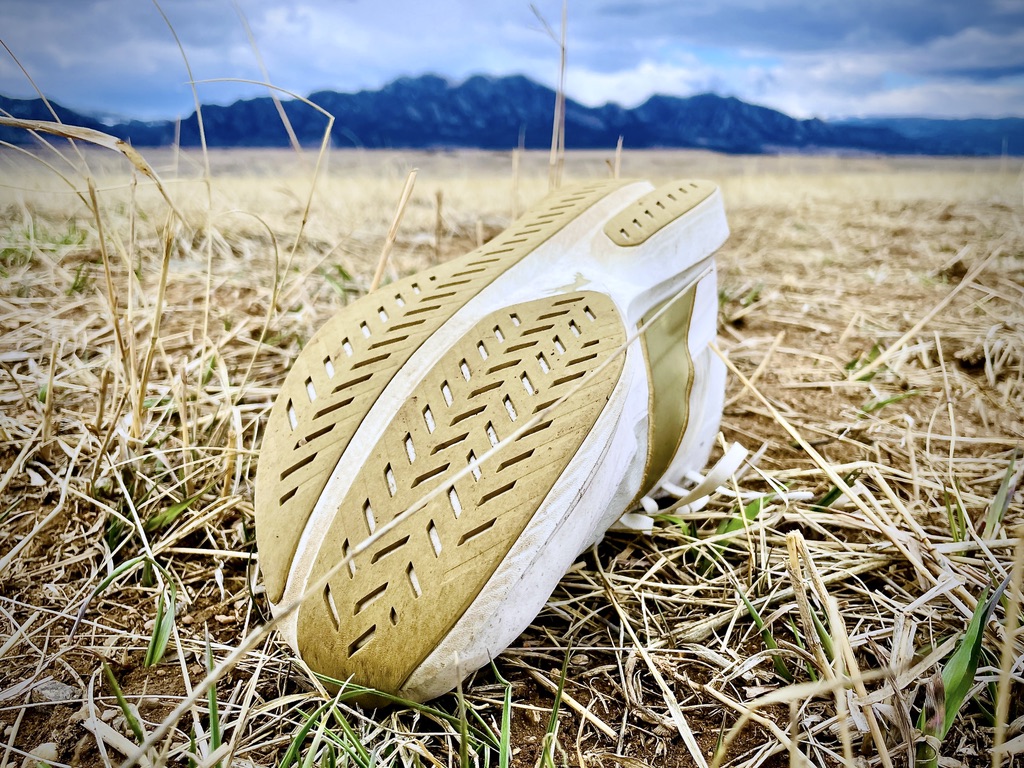 The textured sole of the Tracksmith Eliot Racer rests on dry grass with mountains in the background