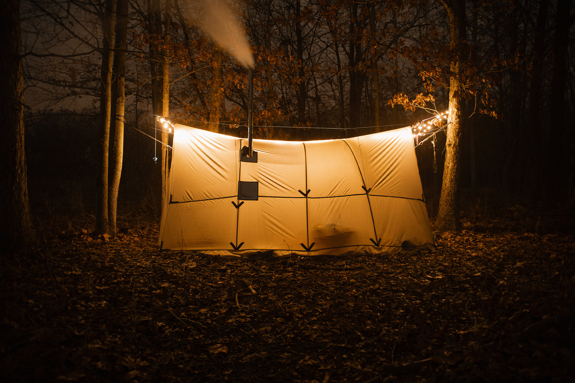 The Warbonnet Tsunami tarp glows warmly at night, with a stove pipe releasing smoke
