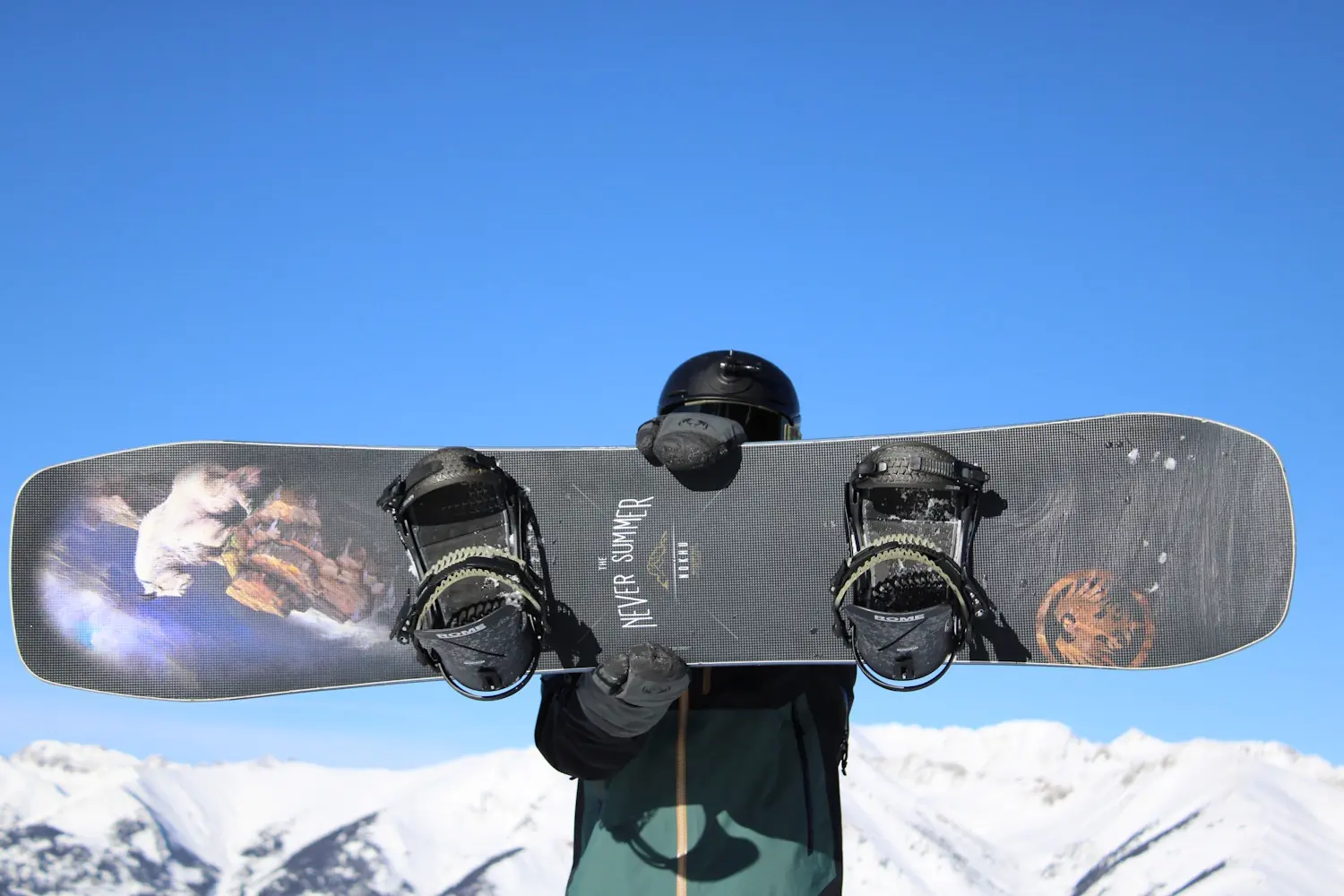 A snowboarder holds up a Never Summer snowboard with Rome Katana bindings