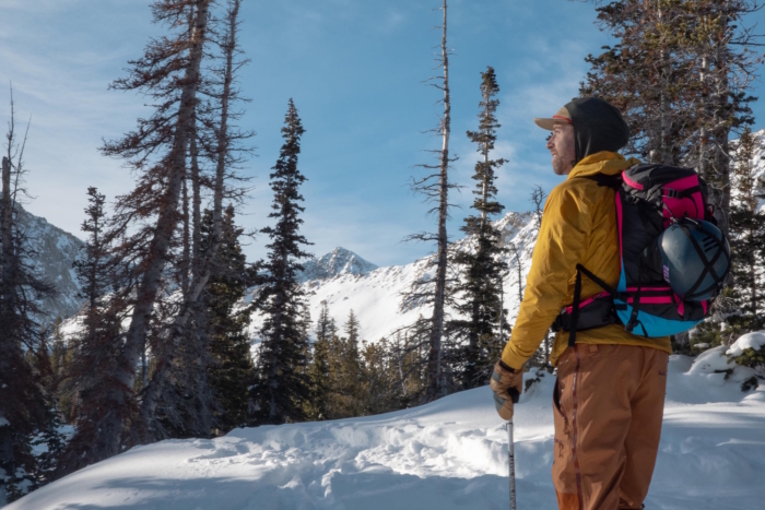 A skier wearing the Pingora Vivid 27L backpack in a snowy backcountry setting, looking toward distant mountain peaks