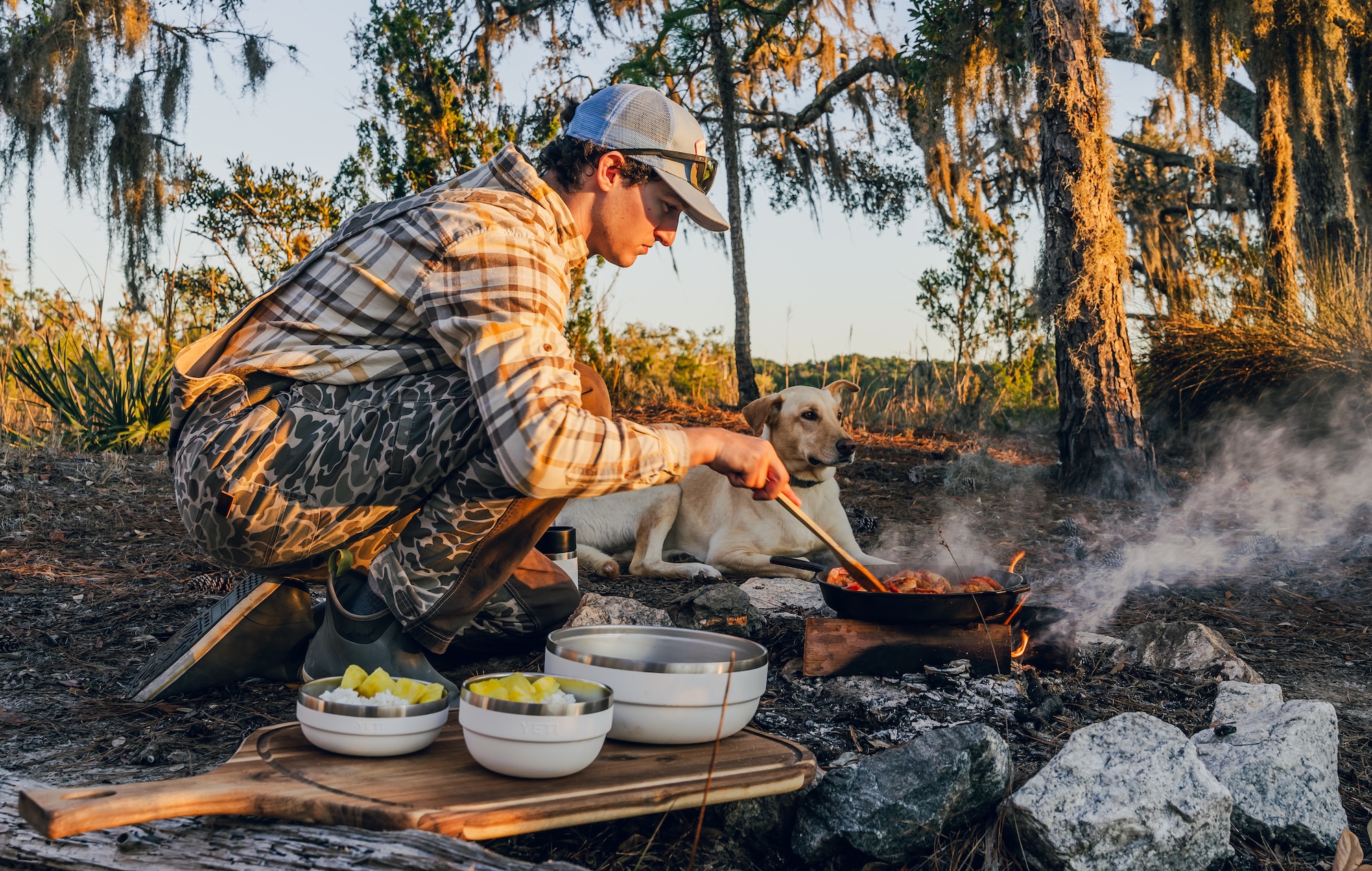 Man and dog cooking on campfire with Yeti rambler bowls next to them