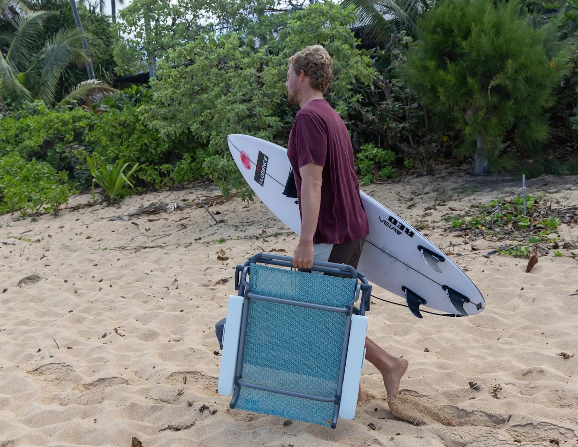 man carrying surfboard and beach chair across sand