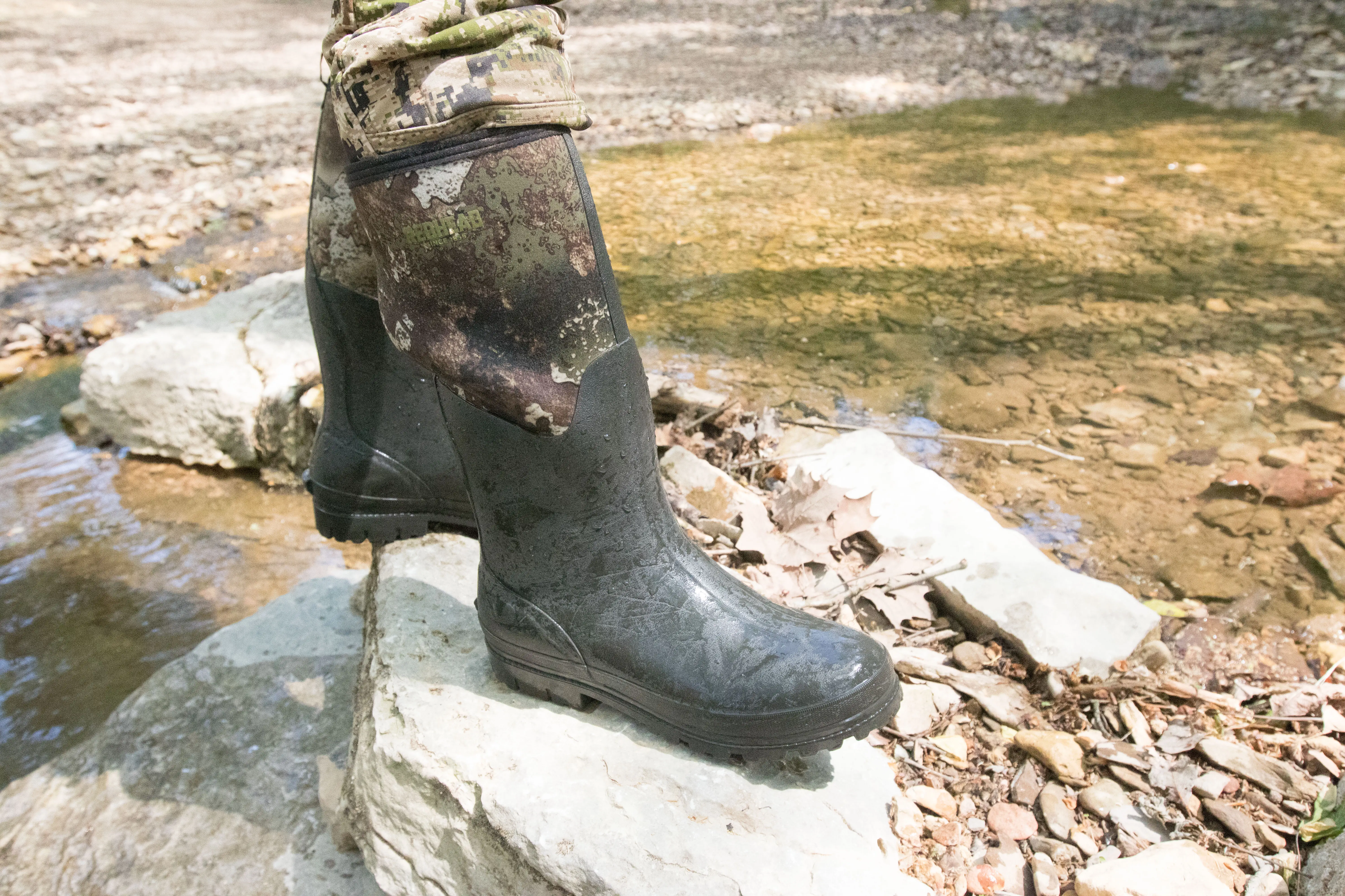 A hunter in rubber boots standing on a rock by a creek.