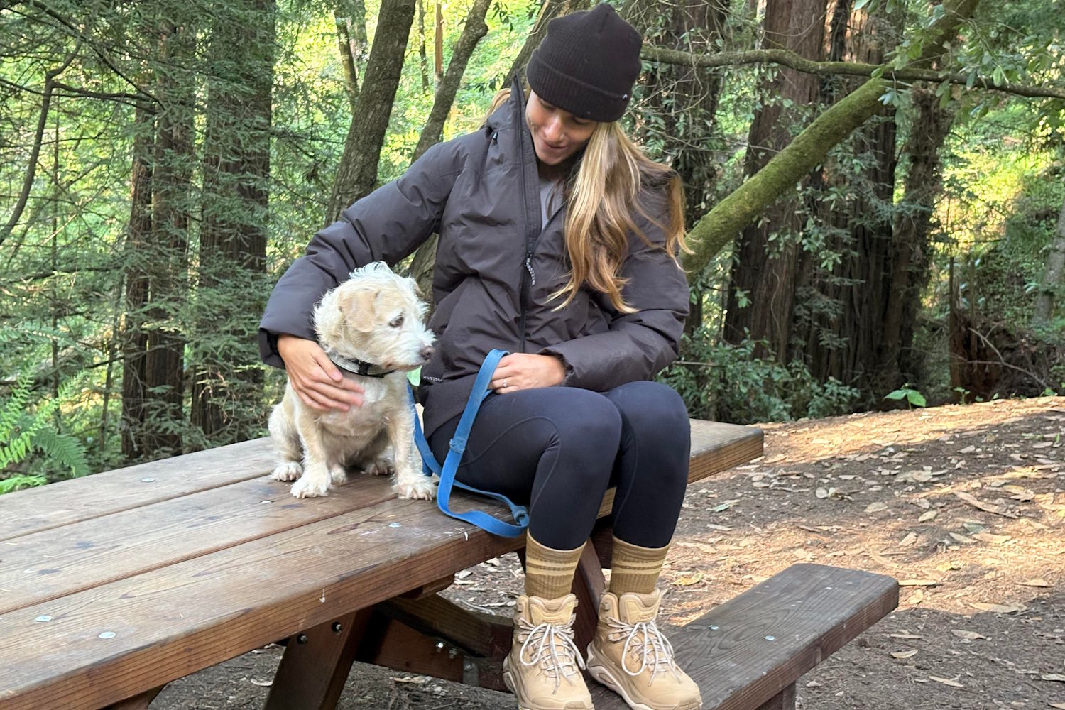 Person sitting on a bench in the forest, wearing a Patagonia jacket and petting a small dog on a leash