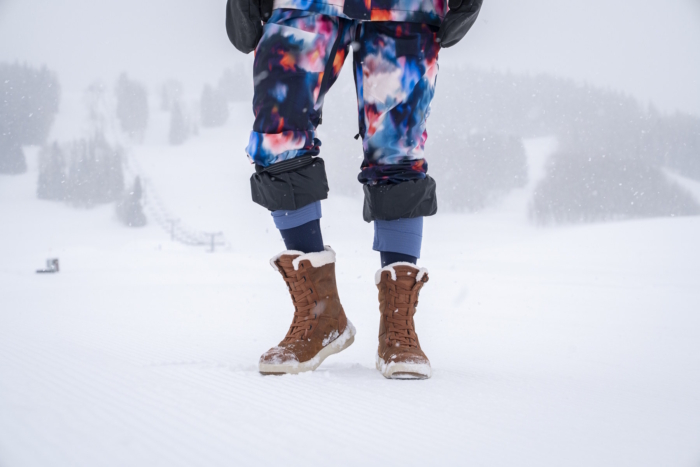Person standing in snow wearing brown Stio Colter boots and colorful snow gear.