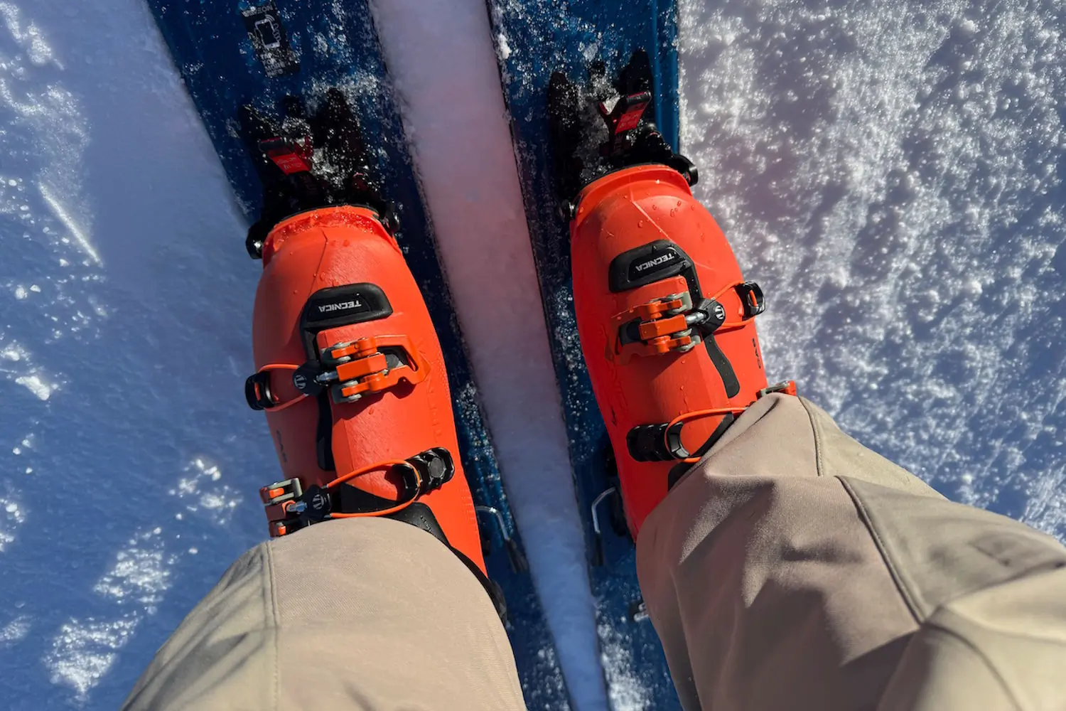 Top-down view of a skier's feet wearing bright orange Tecnica ski boots clipped into blue skis 