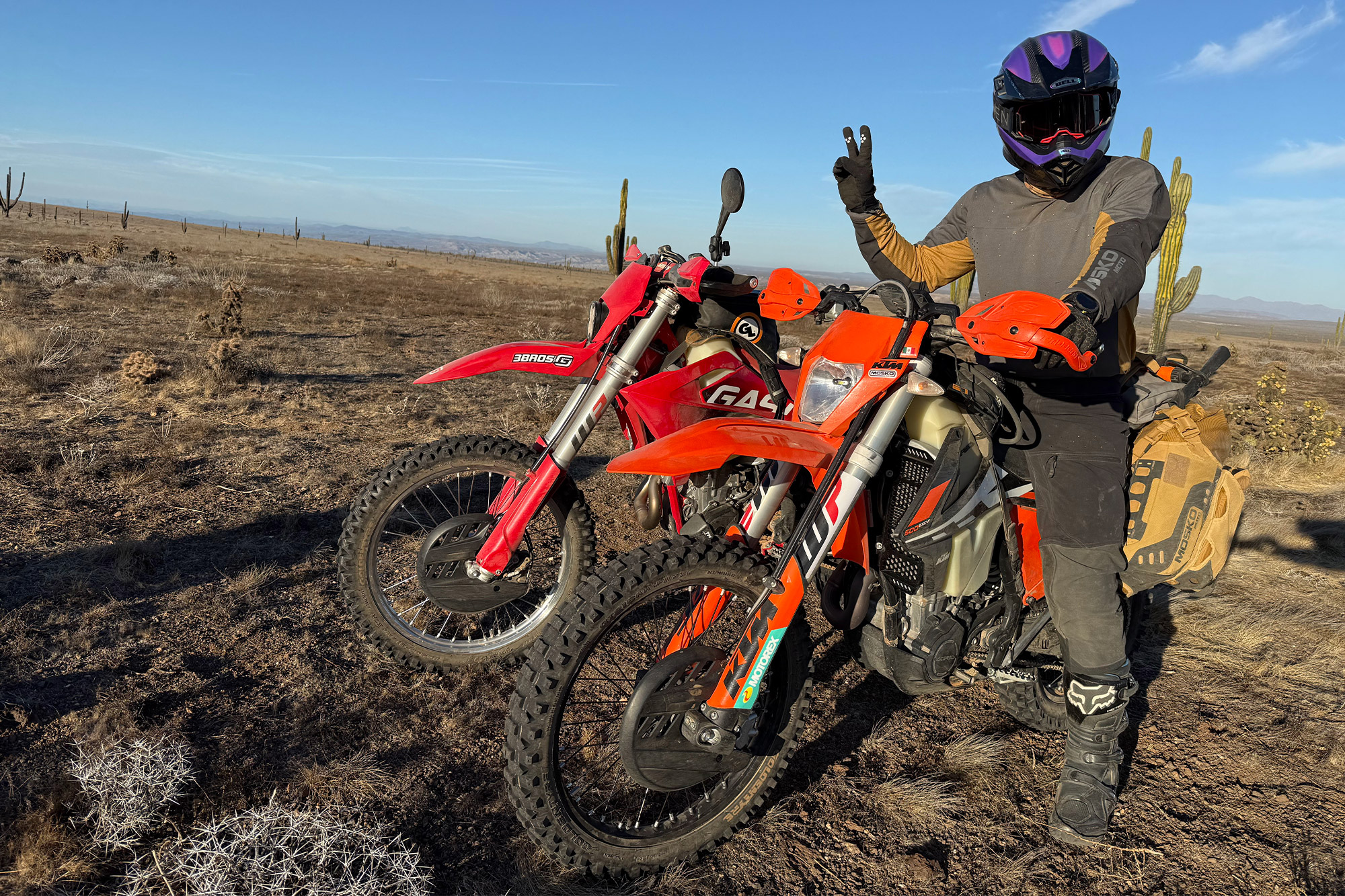 Mosko Moto Reckless 40L System V4.0 strapped to a KTM dirt bike parked in the desert with a rider flashing a peace sign
