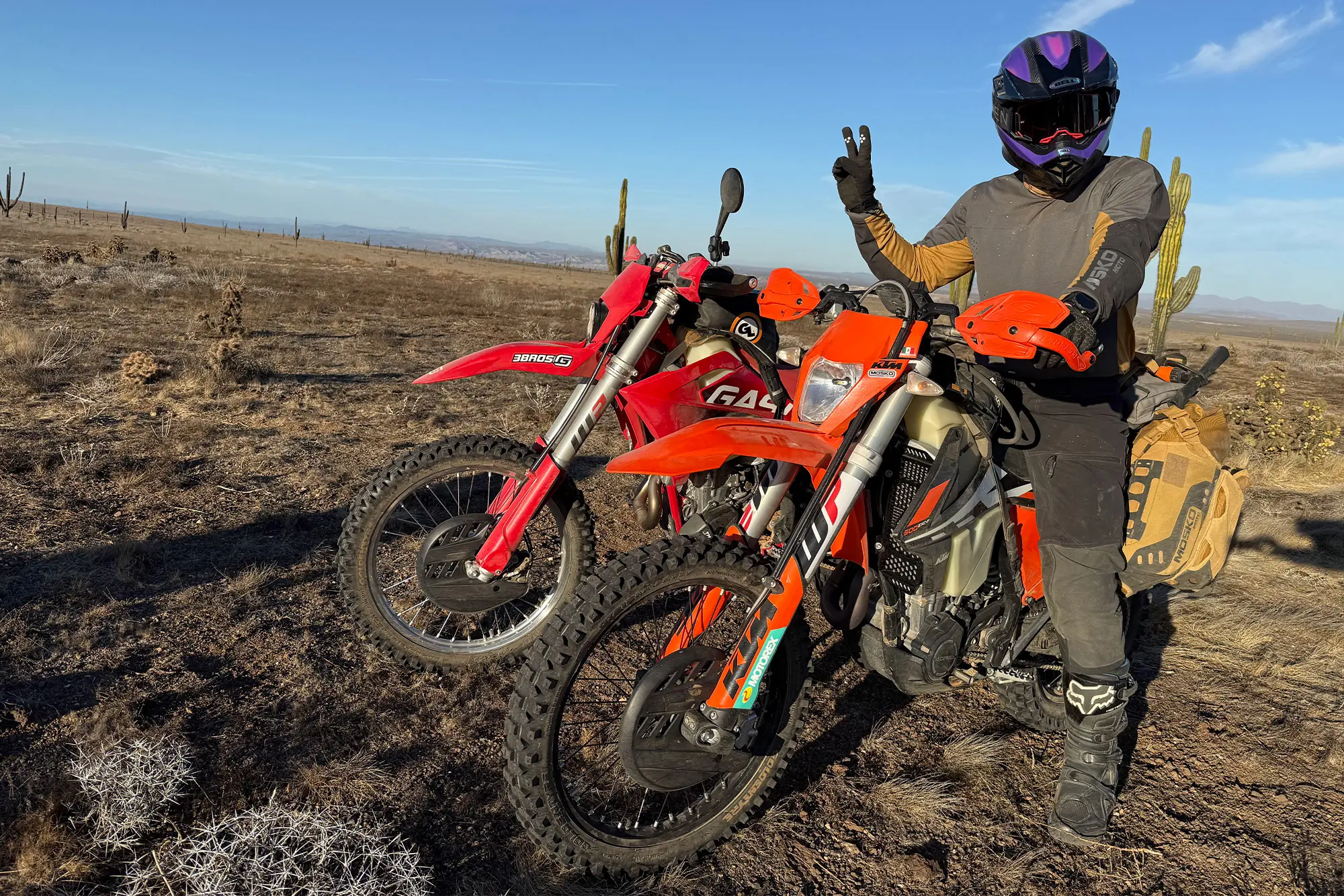 Mosko Moto Reckless 40L System V4.0 strapped to a KTM dirt bike parked in the desert with a rider flashing a peace sign