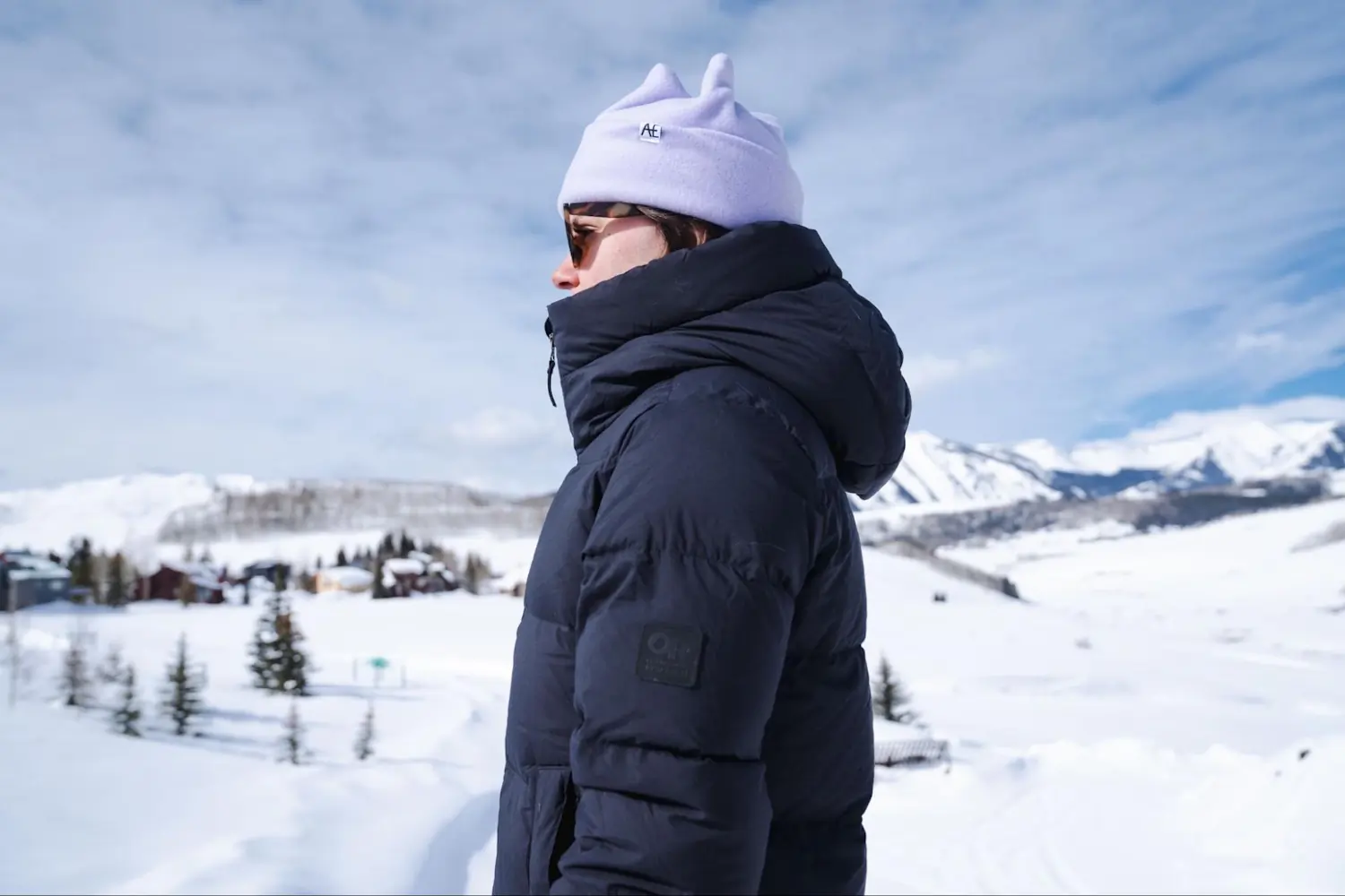 Side view of a woman standing in the snow, wearing a purple beanie and an Outdoor Research Coze Down Parka