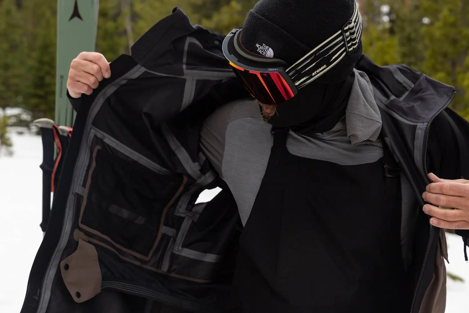 A person wearing ski gear and goggles opens a black TNF Dragline jacket to show its inner lining and pockets