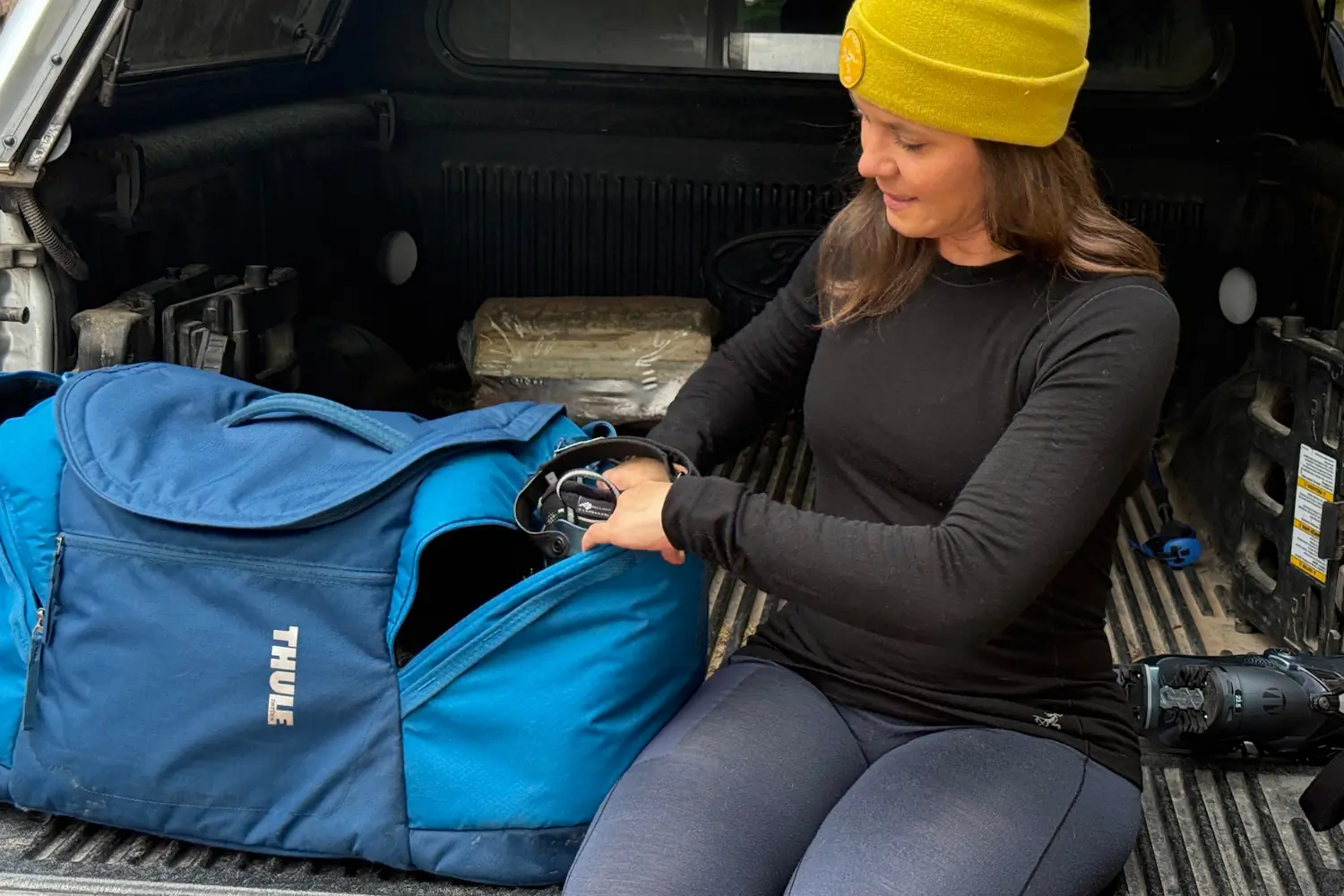 Woman wearing Arc'teryx Satoro base layers packs ski goggles into a blue Thule duffel bag in a truck bed