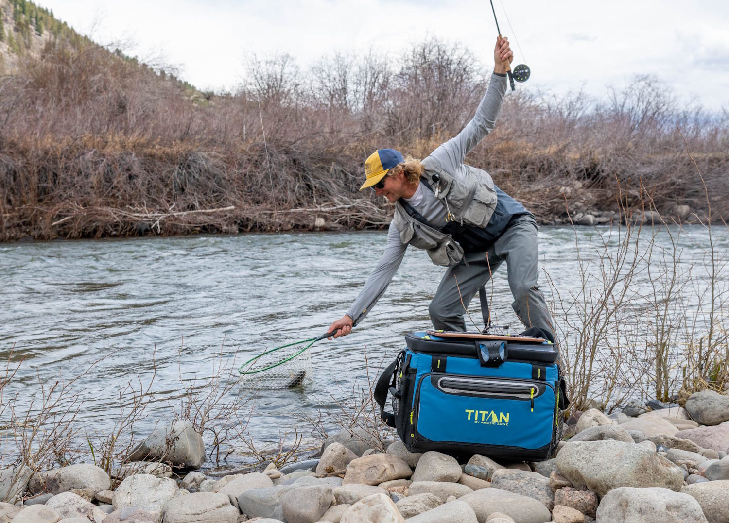 Person fly fishing by a riverbank with a Titan by Arctic Zone 30-can cooler on the rocks nearby.