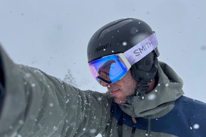 Person wearing a black ski helmet and Smith ski goggles in snowy conditions, dressed in a gray and navy jacket.