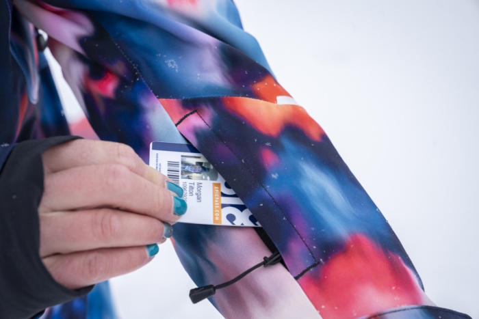 Close-up of a hand inserting a ski pass into the zippered sleeve pocket on the left forearm of a Burton snow jacket.
