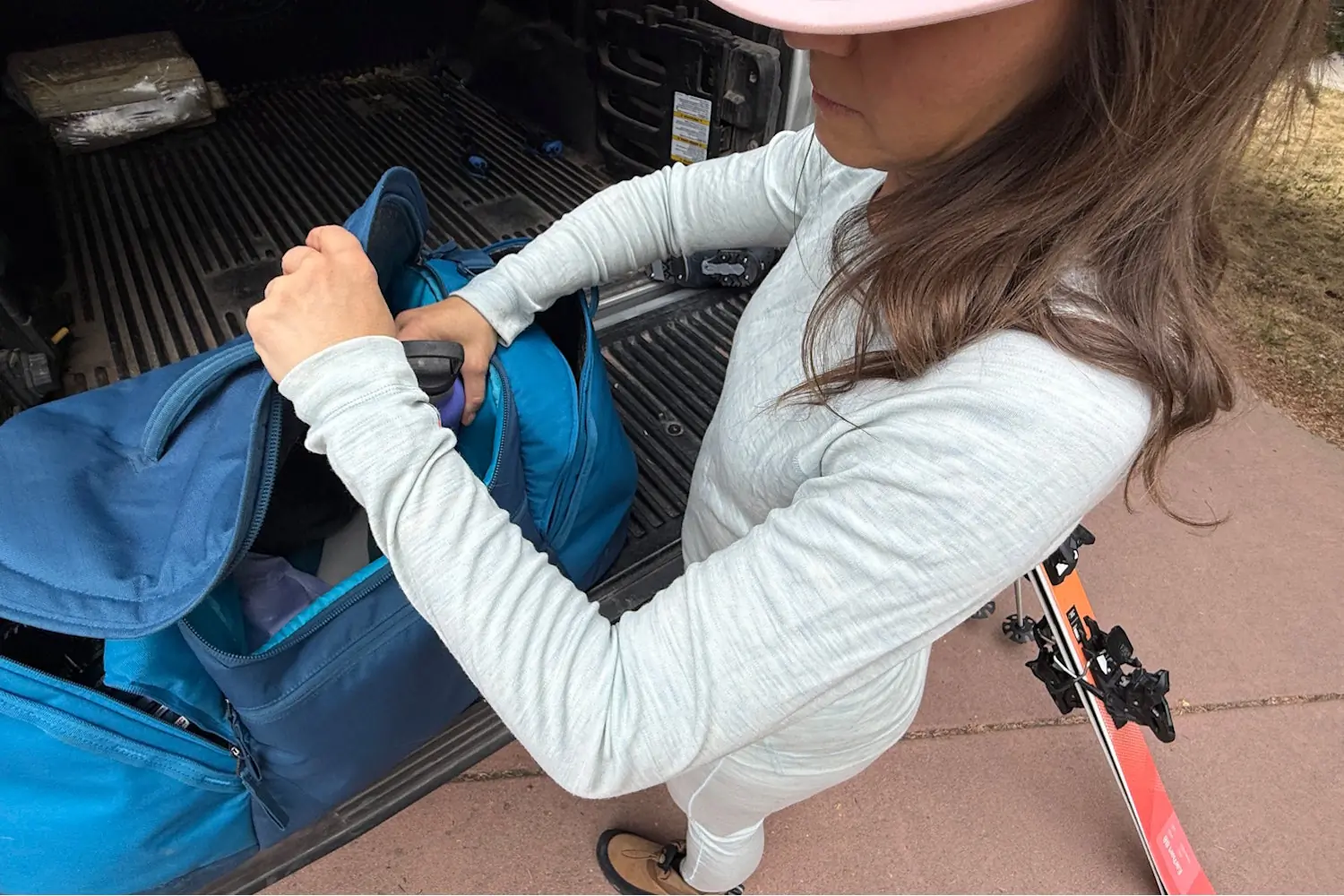 Woman in base layers packs ski gear into a blue duffel next to a truck.