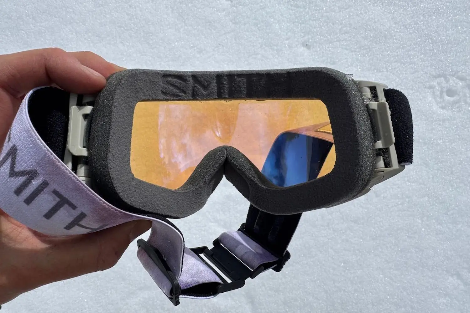 Hand holding a pair of Smith I/O MAG ski goggles with orange lens, shown against a snowy background.