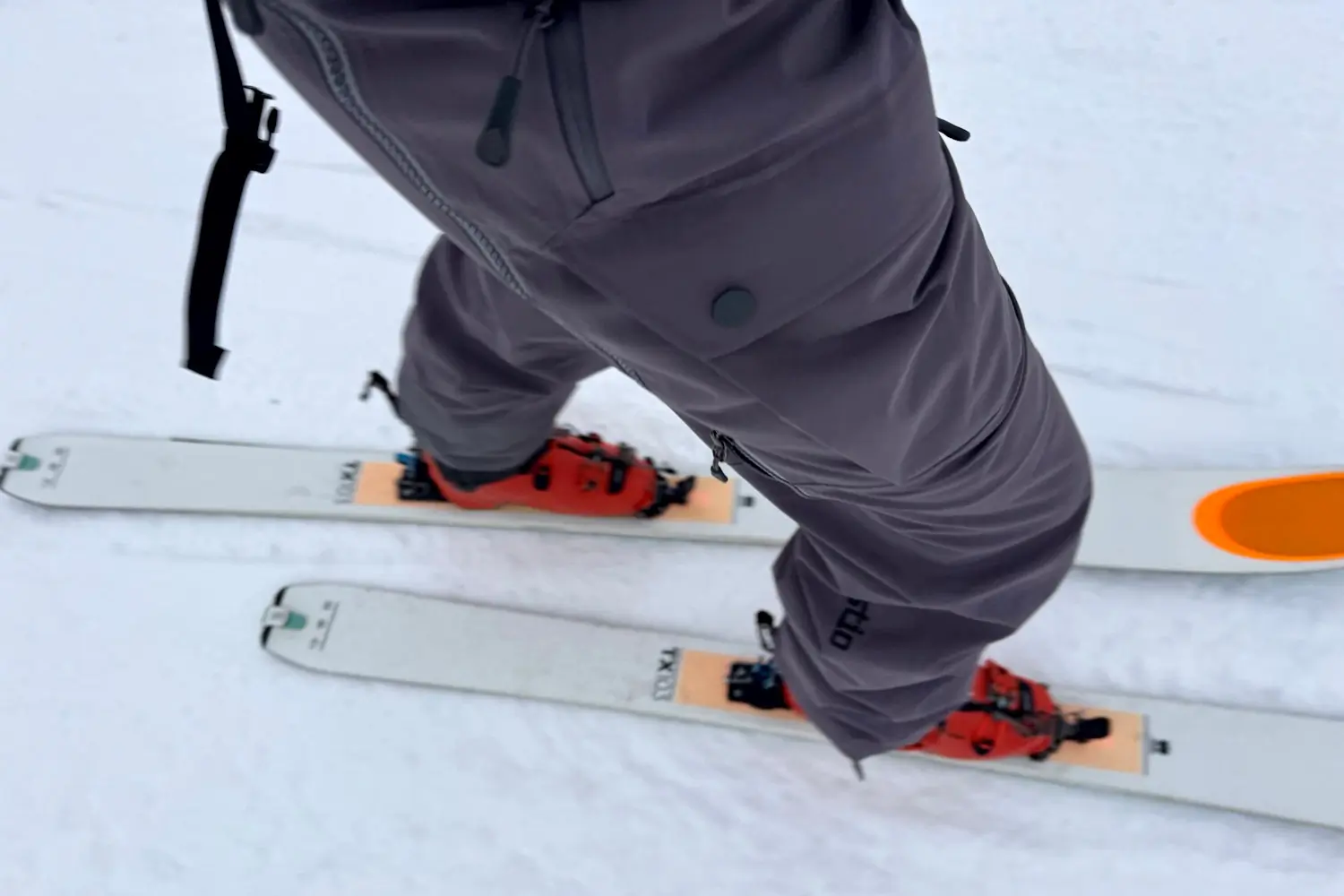 Skier wearing gray Stio Environ pants and red boots on white skis.