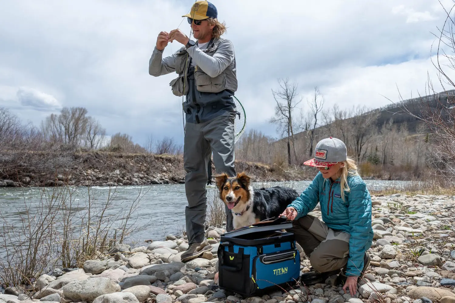 Two people and a dog by a river with a blue Titan cooler; one is fishing, the other opens the cooler