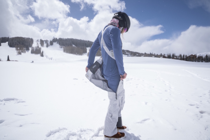 Women adjusts a partially unzipped pair of white Stio Environ Women's Ski Bibs in a snowy mountain landscape