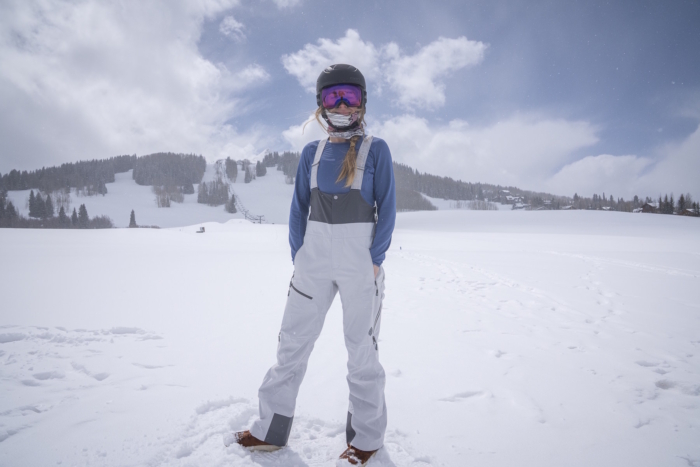 Woman in ski helmet, goggles, and bib overalls standing in a snowy mountain landscape.