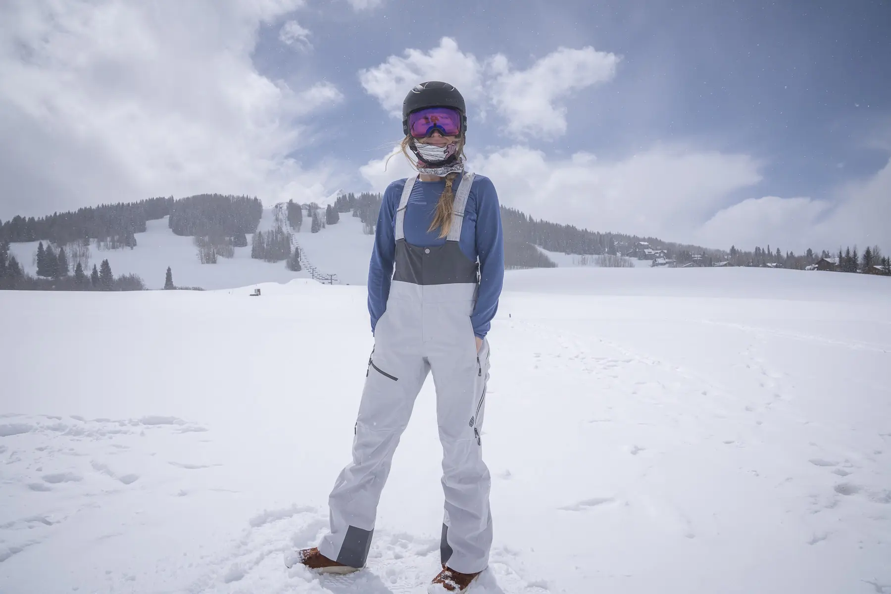 Woman in ski helmet, goggles, and bib overalls standing in a snowy mountain landscape.