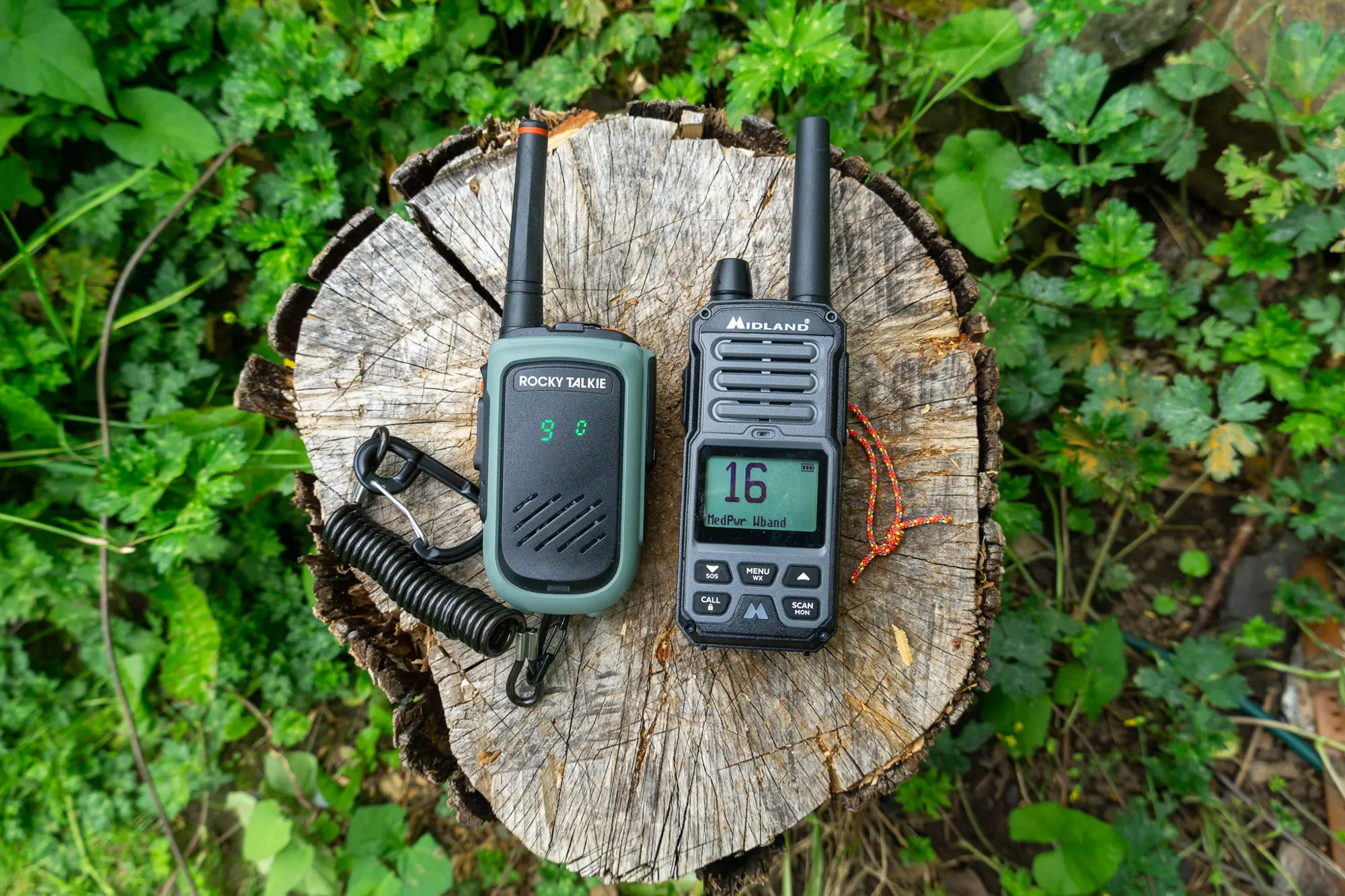 the gxt67 pro and the rocky talkie 5-watt radio side side by side on a stump with a green plant background